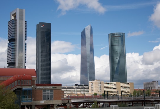 A view of four modern skyscrapers standing tall against a clear blue sky with some clouds. The buildings have a sleek, glassy appearance with varying architectural designs. In the foreground, there's a train station with the sign 'Madrid Chamart&iacute;n', and some residential buildings, trees, and a railway.
