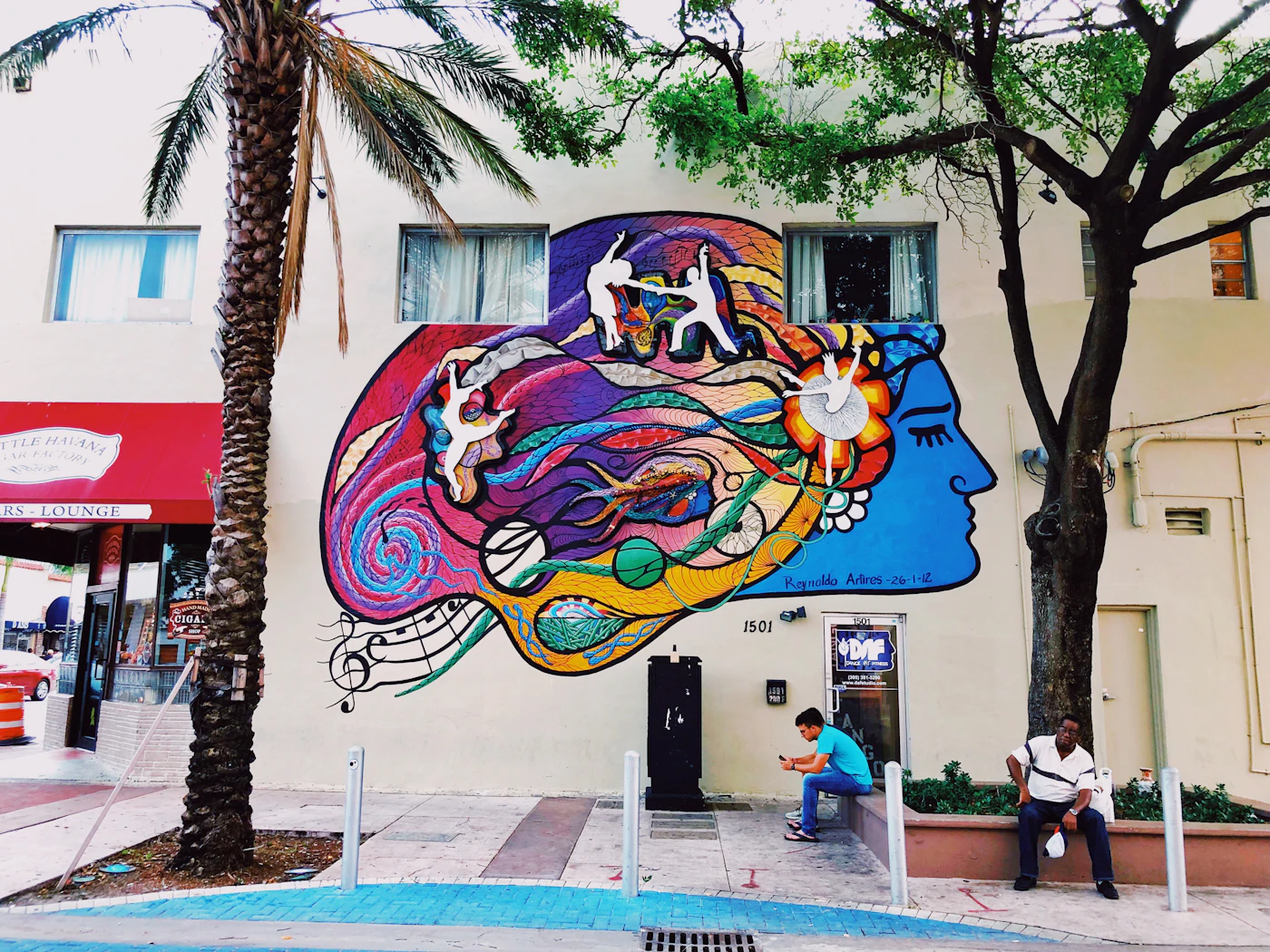 A colorful mural on a Little Havana street corner, with neighbors resting in the shade.