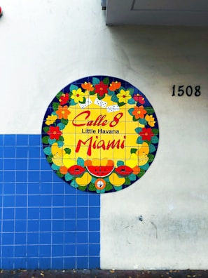 A circular mural on a tiled wall features vibrant floral and fruit designs with dominoes. The center includes 'Calle 8 Little Havana Miami' in bold red letters. The wall is partially covered with blue square tiles and numbered 1508.