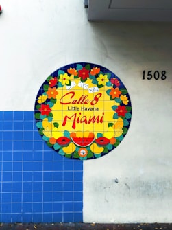 A circular mural on a tiled wall features vibrant floral and fruit designs with dominoes. The center includes 'Calle 8 Little Havana Miami' in bold red letters. The wall is partially covered with blue square tiles and numbered 1508.