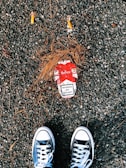 A pair of black and white sneakers are positioned on a rough, gravel-covered ground. Between the shoes lies a red and white cigarette pack with a visible health warning that reads 'Smokers die younger.' Two cigarette butts and scattered pine needles surround the pack.
