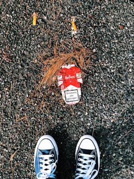 A pair of black and white sneakers are positioned on a rough, gravel-covered ground. Between the shoes lies a red and white cigarette pack with a visible health warning that reads 'Smokers die younger.' Two cigarette butts and scattered pine needles surround the pack.