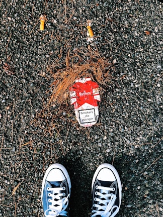 A pair of black and white sneakers are positioned on a rough, gravel-covered ground. Between the shoes lies a red and white cigarette pack with a visible health warning that reads 'Smokers die younger.' Two cigarette butts and scattered pine needles surround the pack.
