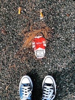 A pair of black and white sneakers are positioned on a rough, gravel-covered ground. Between the shoes lies a red and white cigarette pack with a visible health warning that reads 'Smokers die younger.' Two cigarette butts and scattered pine needles surround the pack.