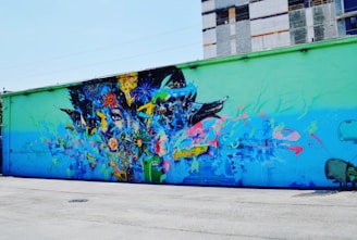 A vibrant street mural covers the side of a building, depicting an abstract composition with elements of nature and technology blended together. Colorful flowers, animal motifs, and machine-like structures merge seamlessly, creating an intricate and dynamic visual tapestry.