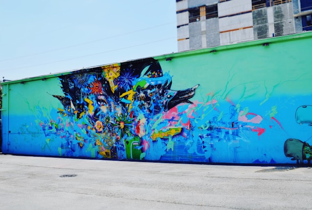A vibrant street mural covers the side of a building, depicting an abstract composition with elements of nature and technology blended together. Colorful flowers, animal motifs, and machine-like structures merge seamlessly, creating an intricate and dynamic visual tapestry.