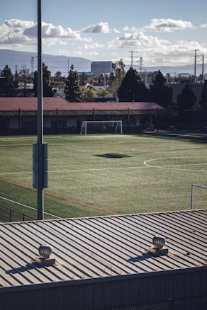 An empty soccer field with green artificial turf is visible, surrounded by a chain-link fence. A metal goalpost stands at one end, and a red-roofed building borders the field. In the background, there are trees, power lines, and a few buildings under a partly cloudy sky. The foreground shows the top of a metal structure with two ventilation units.