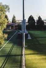 A sports field with artificial turf, divided by a tall pole running down the center. The field is surrounded by trees and buildings in the background, including a structure with a red roof. Shadows play across the grass, creating a contrast between light and shaded areas.