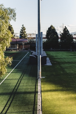 A sports field with artificial turf, divided by a tall pole running down the center. The field is surrounded by trees and buildings in the background, including a structure with a red roof. Shadows play across the grass, creating a contrast between light and shaded areas.