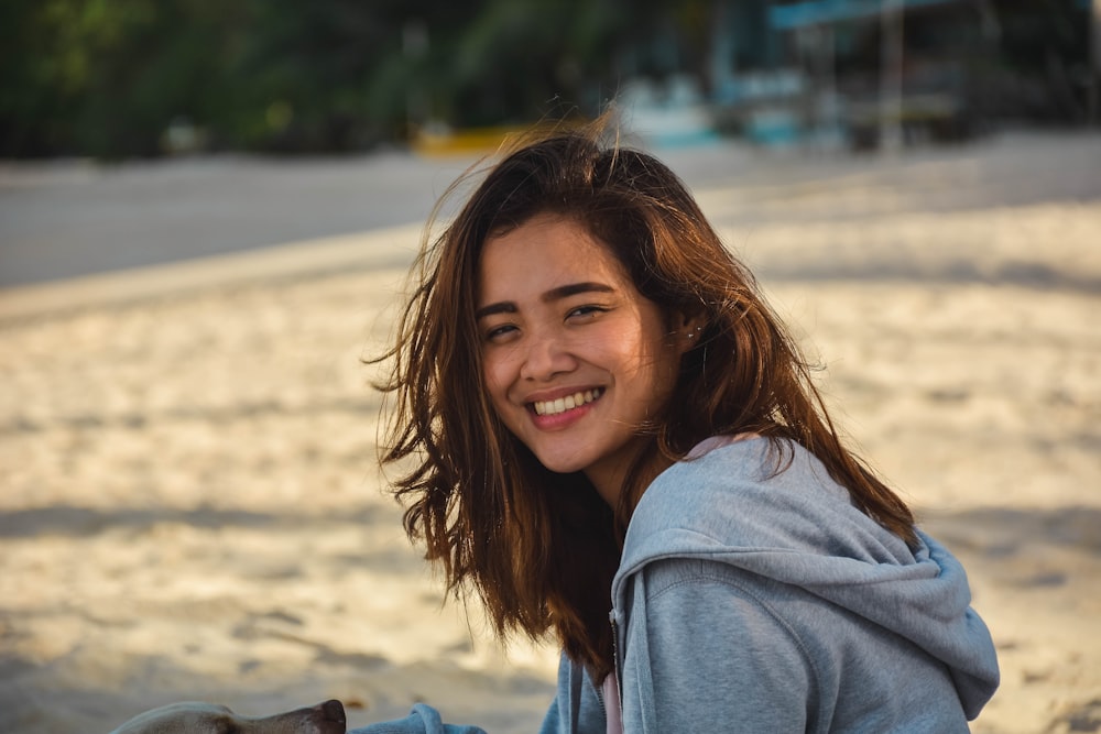woman wearing grey hoodie while sitting on beach sand and smiling