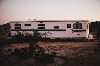 A white, vintage trailer sits in a barren desert landscape. The trailer has several small windows and a door with a curtain hanging inside. Around the trailer are sparse desert plants, and the ground is sandy. The sky above has soft hues, suggesting either dawn or dusk.
