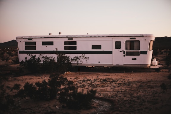 A white, vintage trailer sits in a barren desert landscape. The trailer has several small windows and a door with a curtain hanging inside. Around the trailer are sparse desert plants, and the ground is sandy. The sky above has soft hues, suggesting either dawn or dusk.
