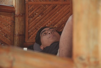 A person wearing glasses is lying on a cushion inside a bamboo structure. The setting appears cozy and relaxed, with the warm tones of wooden bamboo creating a natural and serene environment.