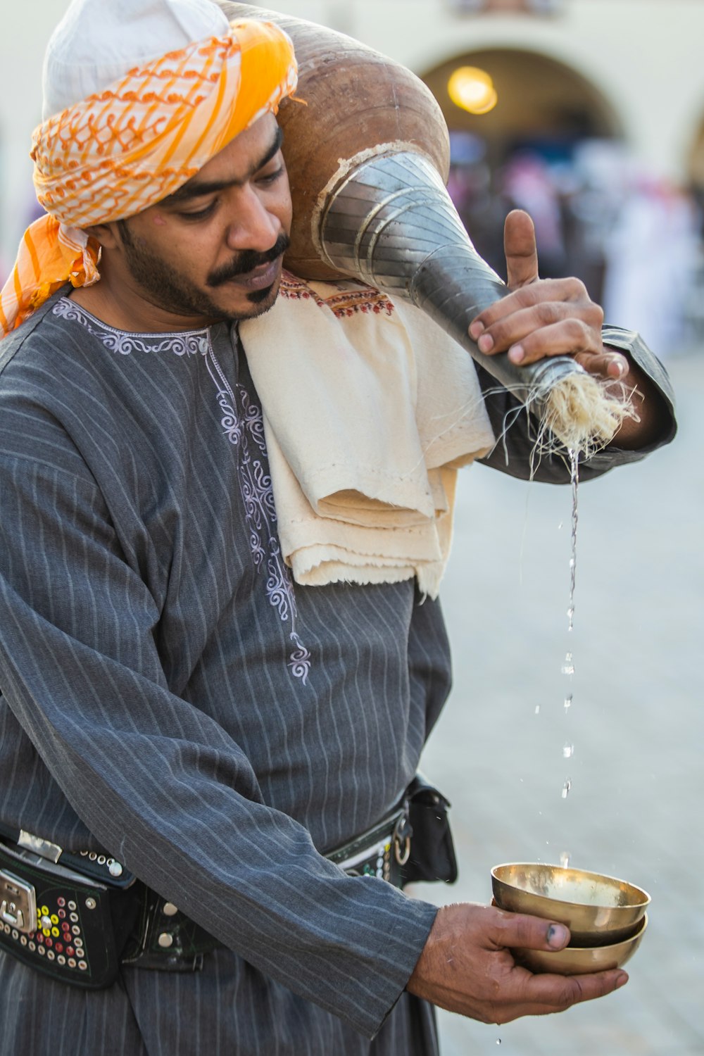 man in grey shirt pouring liquid on brass cup from container on his shoulder