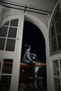 A person sits on the edge of a balcony railing at night, holding a book in their hand. They wear a gray jacket, and the balcony scene is viewed through a partially open, large arched window. The curtains and the dark sky outside frame the composition.