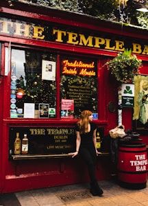 A person with long hair is sitting on a bench in front of a pub with a distinctive red exterior. The pub has signs promoting traditional Irish music and food, with various stickers and posters displayed in the windows. There is a barrel on the right, also marked with the pub's name. Above, there are hanging plants and decorative lights.