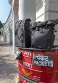 Two black bags are placed on top of a red India Post mailbox. The mailbox displays text in both English and Hindi, indicating sections for letters and packets. The background shows a curved building with white pillars, suggesting an urban setting.