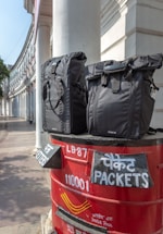Two black bags are placed on top of a red India Post mailbox. The mailbox displays text in both English and Hindi, indicating sections for letters and packets. The background shows a curved building with white pillars, suggesting an urban setting.