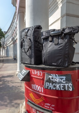 Two black bags are placed on top of a red India Post mailbox. The mailbox displays text in both English and Hindi, indicating sections for letters and packets. The background shows a curved building with white pillars, suggesting an urban setting.
