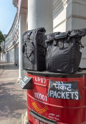 Two black bags are placed on top of a red India Post mailbox. The mailbox displays text in both English and Hindi, indicating sections for letters and packets. The background shows a curved building with white pillars, suggesting an urban setting.
