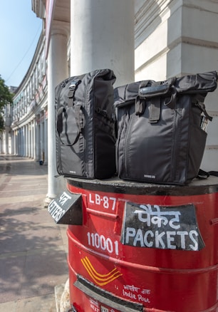 Two black bags are placed on top of a red India Post mailbox. The mailbox displays text in both English and Hindi, indicating sections for letters and packets. The background shows a curved building with white pillars, suggesting an urban setting.