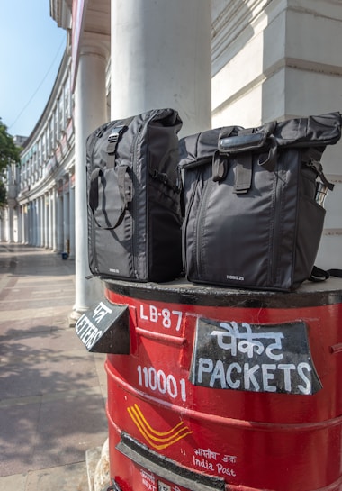Two black bags are placed on top of a red India Post mailbox. The mailbox displays text in both English and Hindi, indicating sections for letters and packets. The background shows a curved building with white pillars, suggesting an urban setting.
