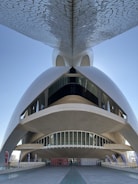 A futuristic and architecturally complex structure with curved forms and a mosaic pattern on the underside of an overhanging element. The building features large, elongated windows and a spacious open area at the base, flanked by promotional banners. The design is both modern and visually striking, set against a clear blue sky.
