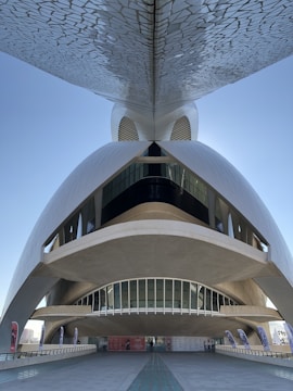 A futuristic and architecturally complex structure with curved forms and a mosaic pattern on the underside of an overhanging element. The building features large, elongated windows and a spacious open area at the base, flanked by promotional banners. The design is both modern and visually striking, set against a clear blue sky.