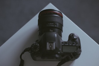 A DSLR camera with a prominent lens sits atop a white surface. The camera is positioned at an angle, showcasing its lens and some dials. The lighting is dim and creates a moody atmosphere.