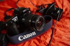 A black Canon DSLR camera placed on a crumpled orange fabric surface. The camera strap is prominently labeled with the Canon logo. Attached cables and a camera charger are visible alongside the camera.