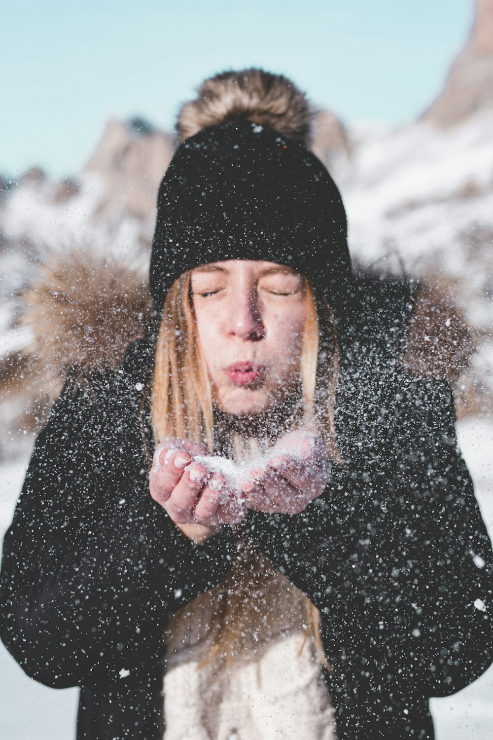 Frau mit schwarzer Bommelmütze und Kapuzenpelzjacke, die Schnee verweht