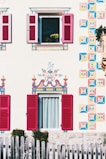 A beautifully decorated house exterior with intricately painted patterns on the facade, featuring vibrant geometric designs and floral motifs around two windows with red shutters. A wooden fence is visible in the foreground, and a flower pot with yellow flowers can be seen through an open window.