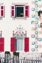 A beautifully decorated house exterior with intricately painted patterns on the facade, featuring vibrant geometric designs and floral motifs around two windows with red shutters. A wooden fence is visible in the foreground, and a flower pot with yellow flowers can be seen through an open window.
