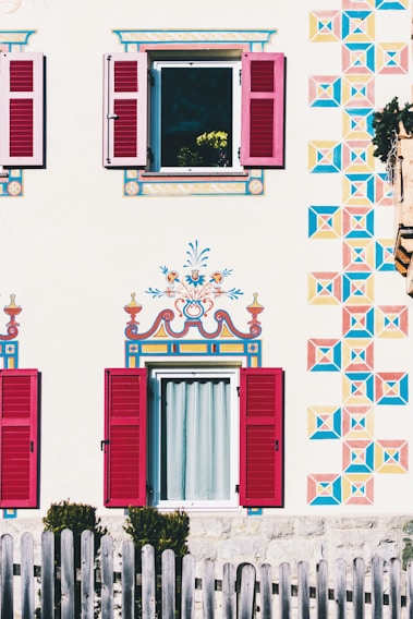 A beautifully decorated house exterior with intricately painted patterns on the facade, featuring vibrant geometric designs and floral motifs around two windows with red shutters. A wooden fence is visible in the foreground, and a flower pot with yellow flowers can be seen through an open window.