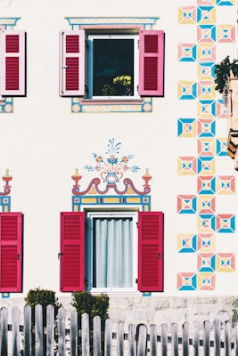 A beautifully decorated house exterior with intricately painted patterns on the facade, featuring vibrant geometric designs and floral motifs around two windows with red shutters. A wooden fence is visible in the foreground, and a flower pot with yellow flowers can be seen through an open window.
