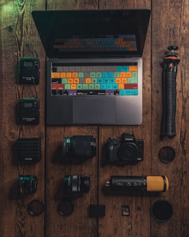 A neatly arranged collection of photography and media equipment is displayed on a rustic wooden surface. In the center, there is a laptop with a colorful keyboard overlay. Surrounding the laptop are various camera lenses, a digital camera, an external microphone with a gold mesh, a tripod, and other small accessories like a memory card and battery pack.