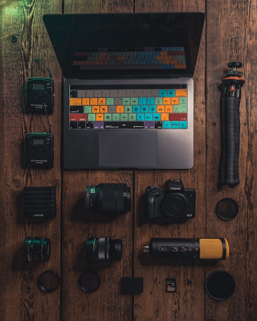 A neatly arranged collection of photography and media equipment is displayed on a rustic wooden surface. In the center, there is a laptop with a colorful keyboard overlay. Surrounding the laptop are various camera lenses, a digital camera, an external microphone with a gold mesh, a tripod, and other small accessories like a memory card and battery pack.
