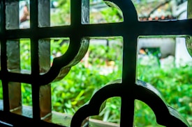 An ornate window grille made of metal is in the foreground, displaying decorative geometric patterns. Through the grille, a lush, green garden is visible in soft focus, hinting at a peaceful and natural outdoor setting.