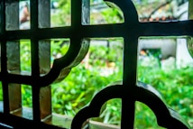 An ornate window grille made of metal is in the foreground, displaying decorative geometric patterns. Through the grille, a lush, green garden is visible in soft focus, hinting at a peaceful and natural outdoor setting.