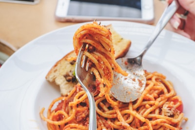 A plate of spaghetti with a tomato-based sauce, being twirled on a fork held above the dish. A slice of bread is placed on the plate, and a spoon assists in twirling the pasta. A smartphone is partially visible in the background on the table.