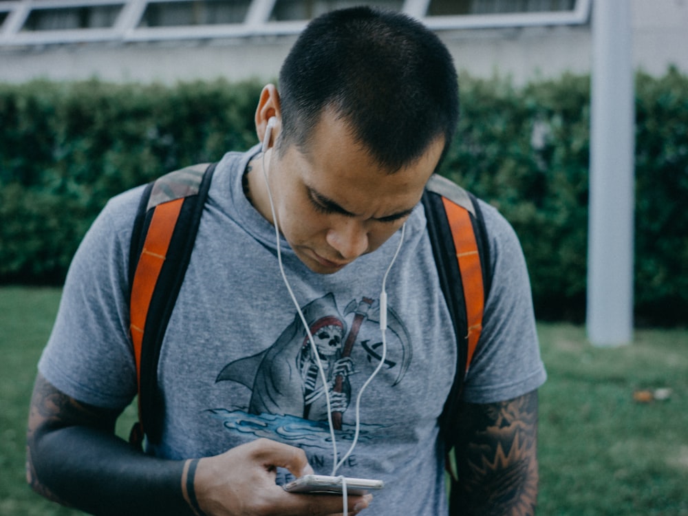 man wearing gray t-shirt, orange backpack, and white earphones while holding phone