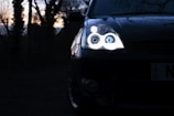 A close-up of a car's headlight shining brightly in a dimly lit environment. The scene features a dark, wooded background with silhouettes of trees at sunset.