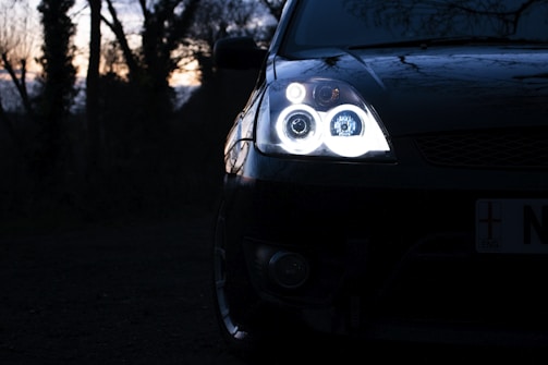 A close-up of a car's headlight shining brightly in a dimly lit environment. The scene features a dark, wooded background with silhouettes of trees at sunset.