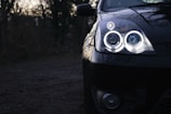 A close-up view of a car's front end highlighting its illuminated headlights with a double halo design. The shiny black surface of the car reflects the surroundings, and the vehicle is parked in a dimly lit outdoor area surrounded by blurred trees.