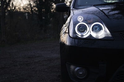 A close-up view of a car's front end highlighting its illuminated headlights with a double halo design. The shiny black surface of the car reflects the surroundings, and the vehicle is parked in a dimly lit outdoor area surrounded by blurred trees.