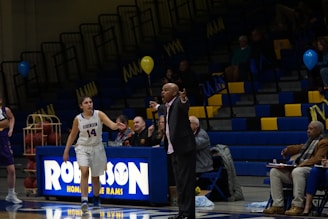 A basketball game is taking place in a gymnasium, focusing on a coach passionately giving directions from the sidelines. A female player in a white jersey is walking on the court, and in the background, the stands are partially filled with spectators. The environment includes balloons and a scoreboard with the word 'Robinson'.