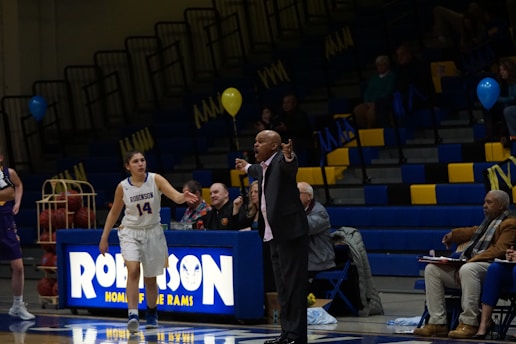 A basketball game is taking place in a gymnasium, focusing on a coach passionately giving directions from the sidelines. A female player in a white jersey is walking on the court, and in the background, the stands are partially filled with spectators. The environment includes balloons and a scoreboard with the word 'Robinson'.
