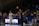A basketball game is taking place in a gymnasium, focusing on a coach passionately giving directions from the sidelines. A female player in a white jersey is walking on the court, and in the background, the stands are partially filled with spectators. The environment includes balloons and a scoreboard with the word 'Robinson'.