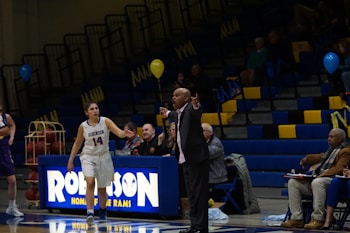A basketball game is taking place in a gymnasium, focusing on a coach passionately giving directions from the sidelines. A female player in a white jersey is walking on the court, and in the background, the stands are partially filled with spectators. The environment includes balloons and a scoreboard with the word 'Robinson'.