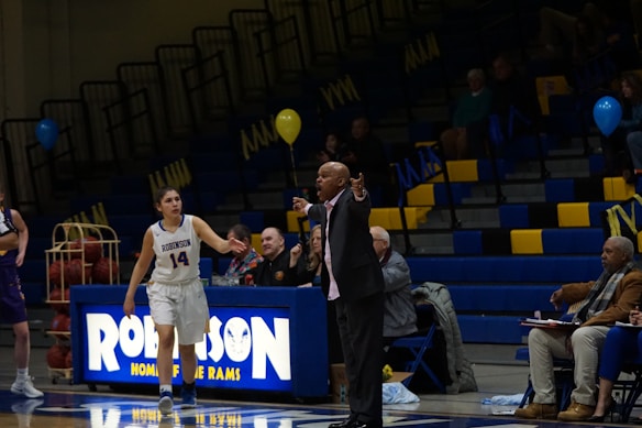 A basketball game is taking place in a gymnasium, focusing on a coach passionately giving directions from the sidelines. A female player in a white jersey is walking on the court, and in the background, the stands are partially filled with spectators. The environment includes balloons and a scoreboard with the word 'Robinson'.